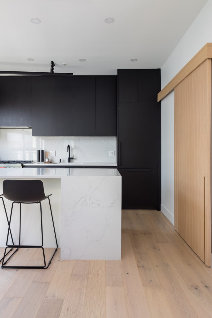 Full view of a contemporary white rock kitchen with matte black upper and integrated lower cabinetry, a white quartz waterfall island with bar seating, and light-colored hardwood floors. Features a custom wood sliding door for access.