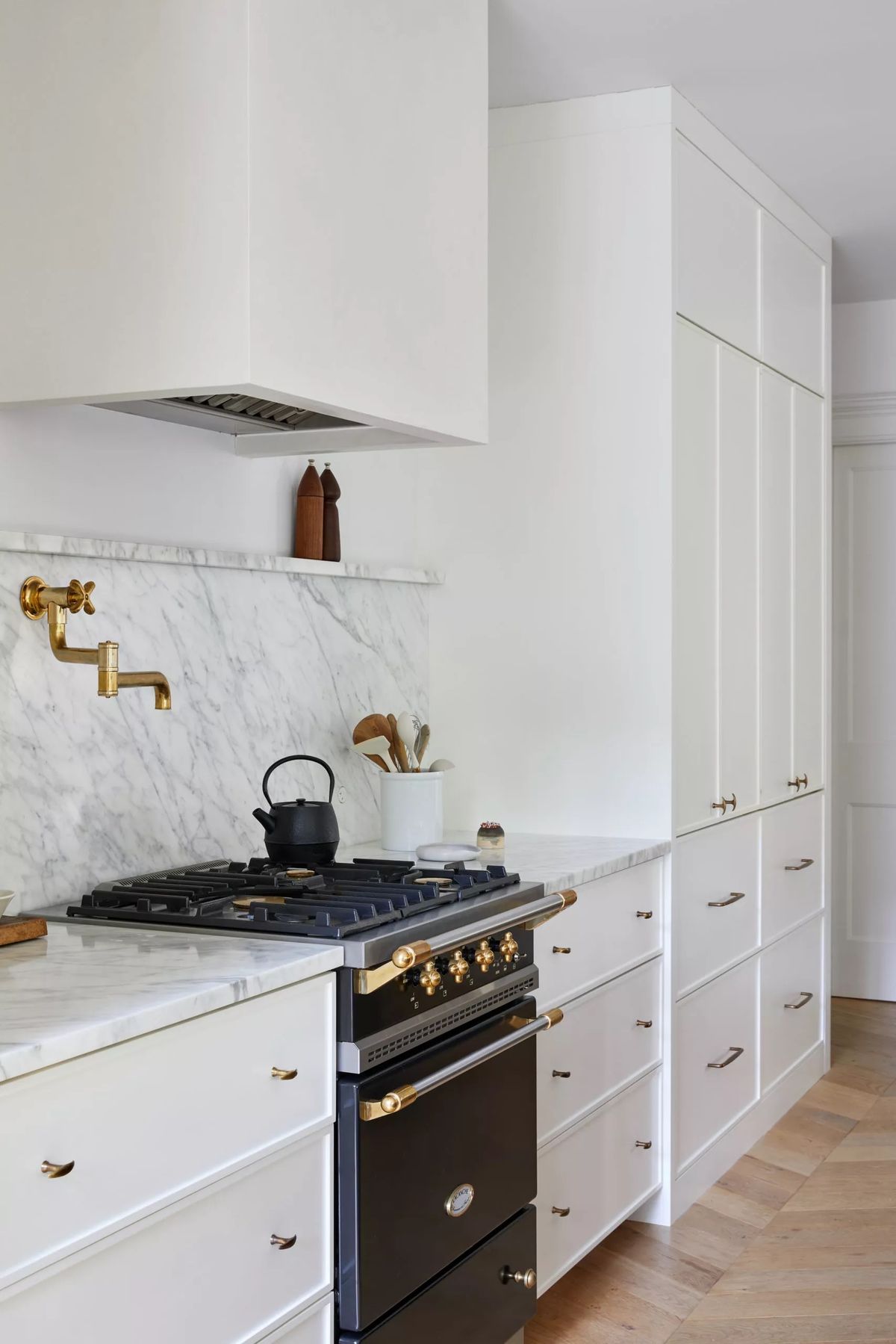 Bright modern kitchen with white cabinetry, marble backsplash, brass pot filler, and a black-gold gas range.