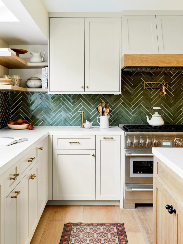 Coquitlam kitchen layout featuring dark green herringbone backsplash tiles, cream shaker cabinets, gold hardware, and open shelving in a functional corner workspace.