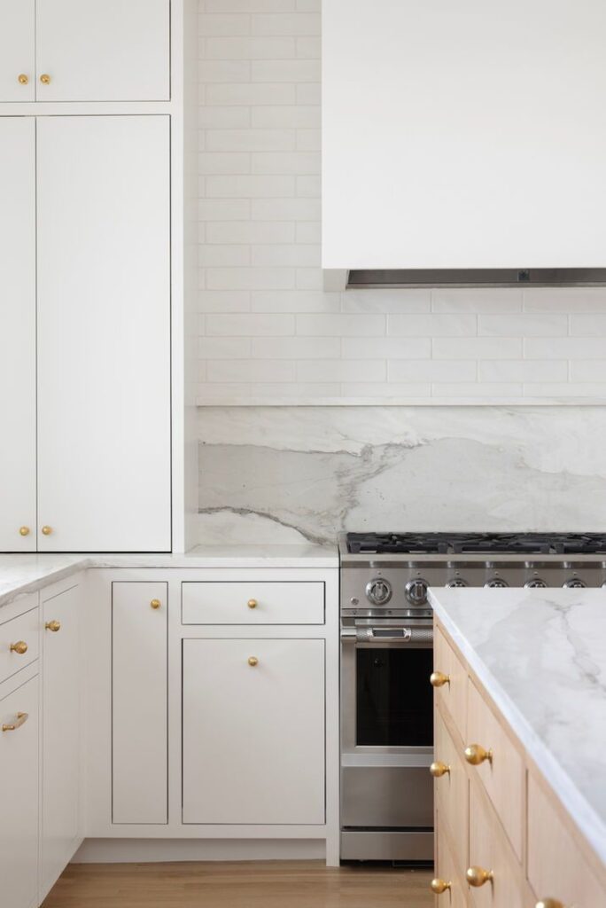 Transitional Coquitlam kitchen design featuring white shaker cabinets, a light wood island with gold knobs, and a mixed material backsplash with a marble slab ledge and white subway tiles.