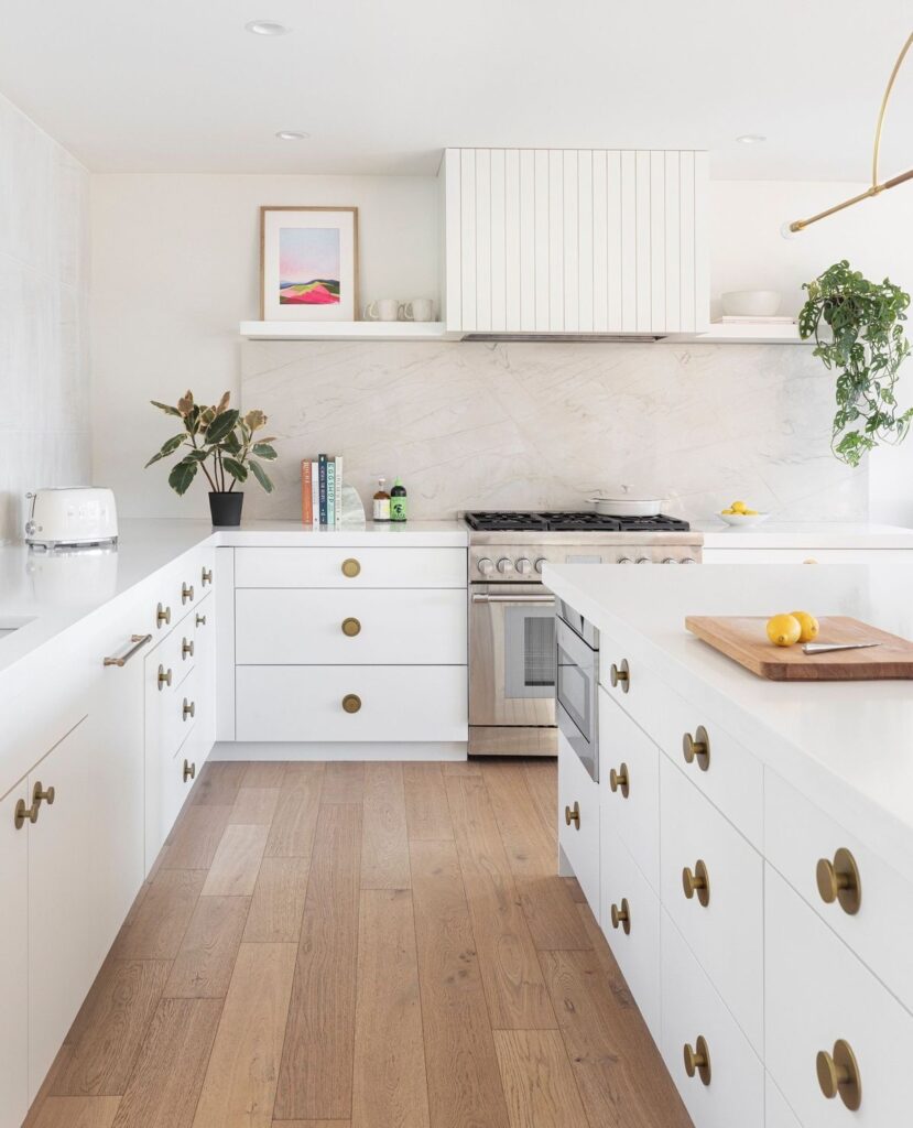 Bright U-shaped kitchen layout in a Coquitlam home featuring white cabinetry with gold round knobs, a paneled range hood, and a stone slab backsplash.