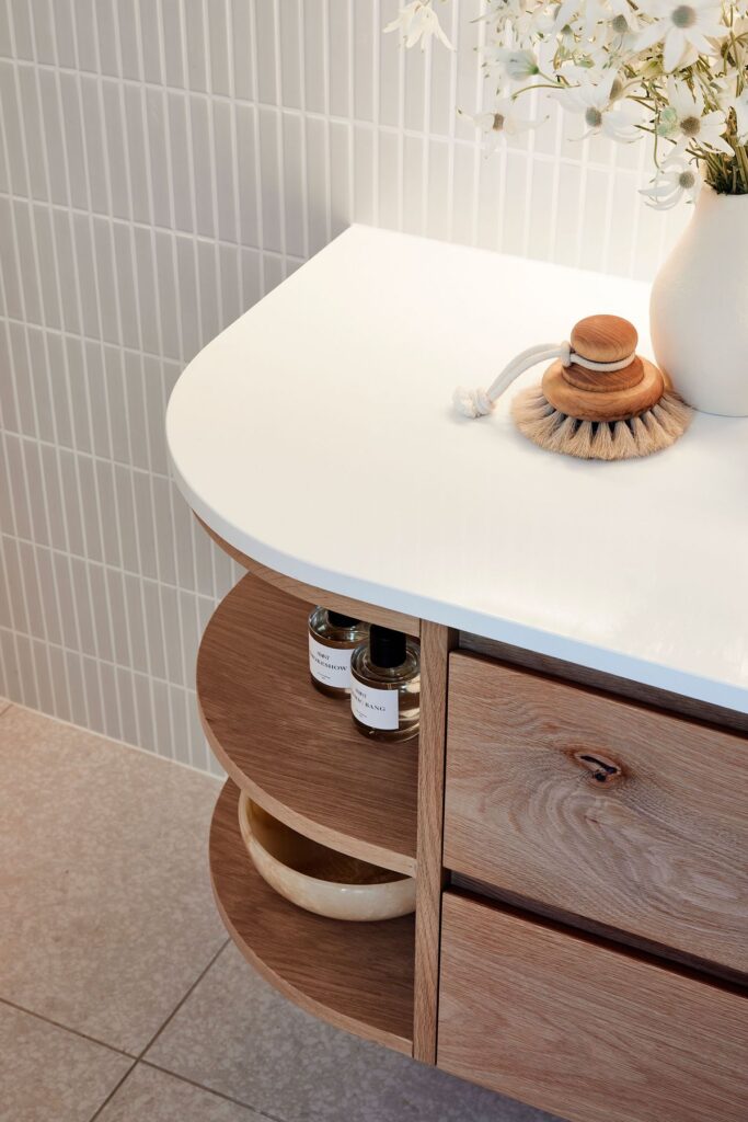 Close-up of a modern bathroom vanity featuring a curved, light oak wood cabinet base with open rounded shelving and a solid white countertop. The counter is styled with a natural wood and bristle brush and white vase.