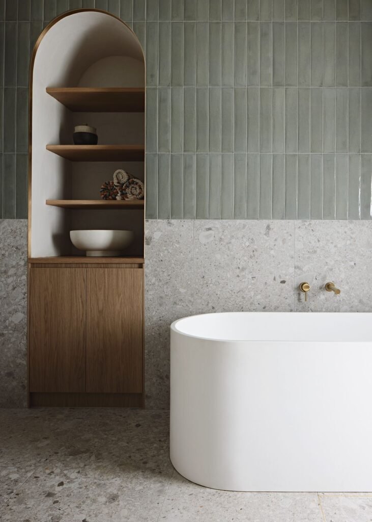 Modern luxury bathroom interior with a white freestanding bathtub, sage green vertical subway tiles, and gray terrazzo flooring and wainscoting. Built-in wood and arch niche shelving with brass trim on the left.