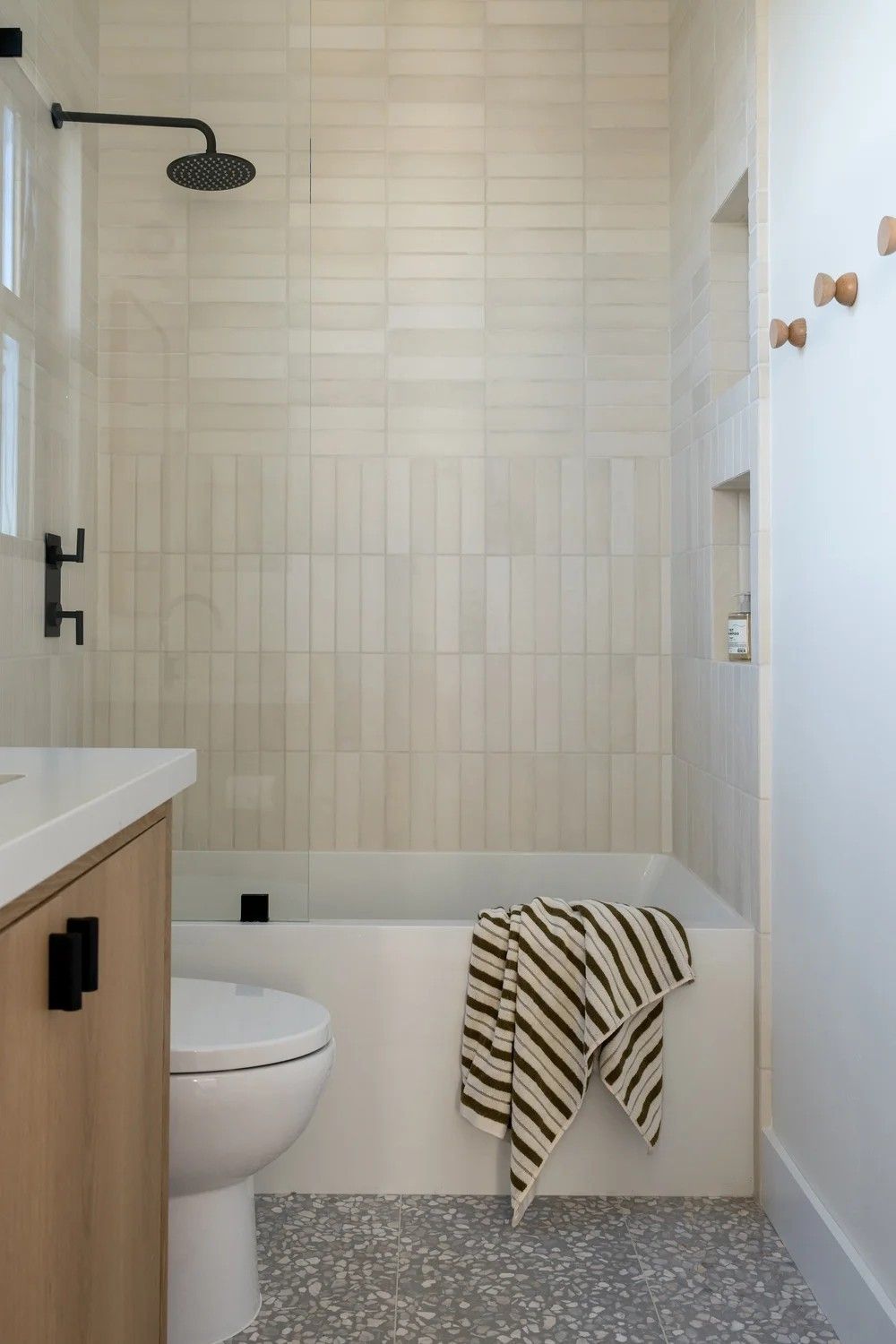 View of a small, modern bathroom featuring a bathtub and shower combination tiled with glossy white vertical subway tiles. Includes a wall niche, matte black shower fixtures, a wooden vanity on the left, and gray speckled floor tiles