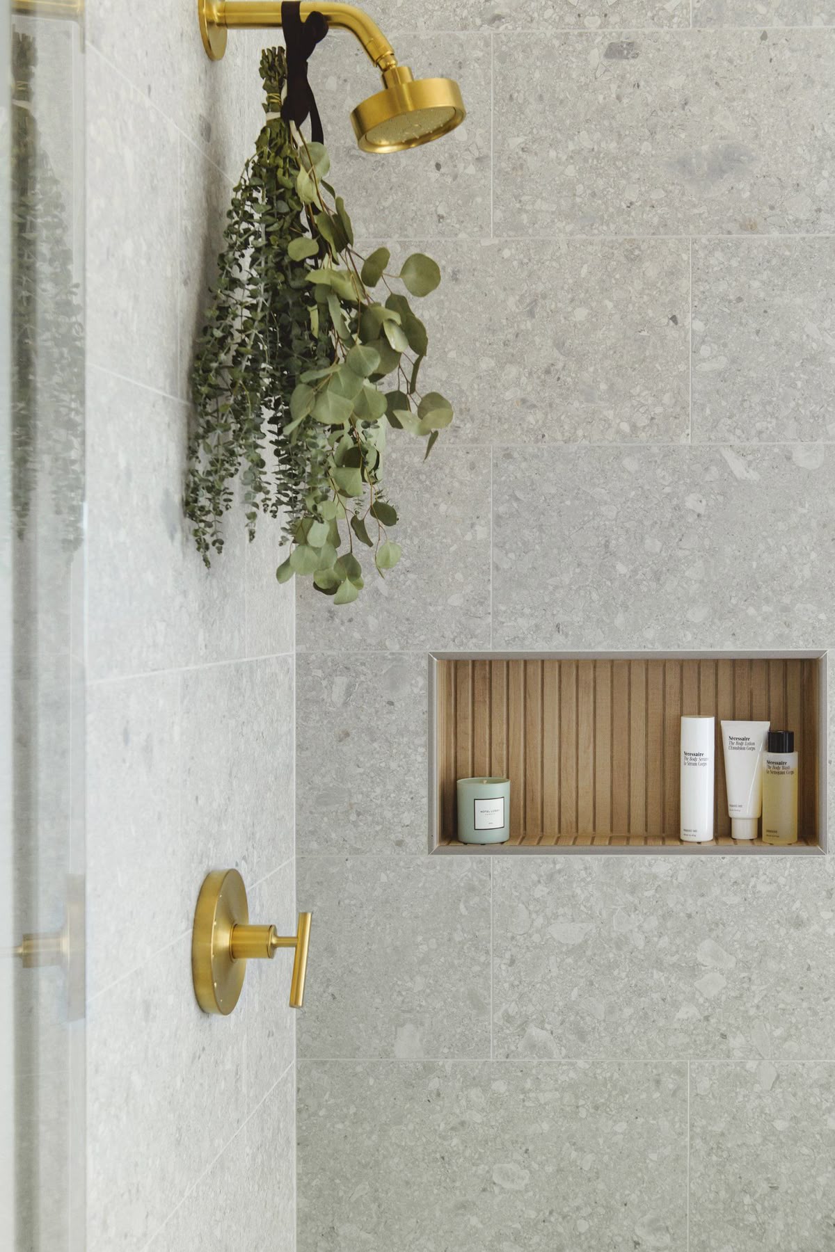Close-up of a shower wall tiled with large-format gray speckled terrazzo, featuring a built-in niche with a slatted wood back. The shower has brushed brass fixtures, and a bunch of eucalyptus is hanging from the showerhead.