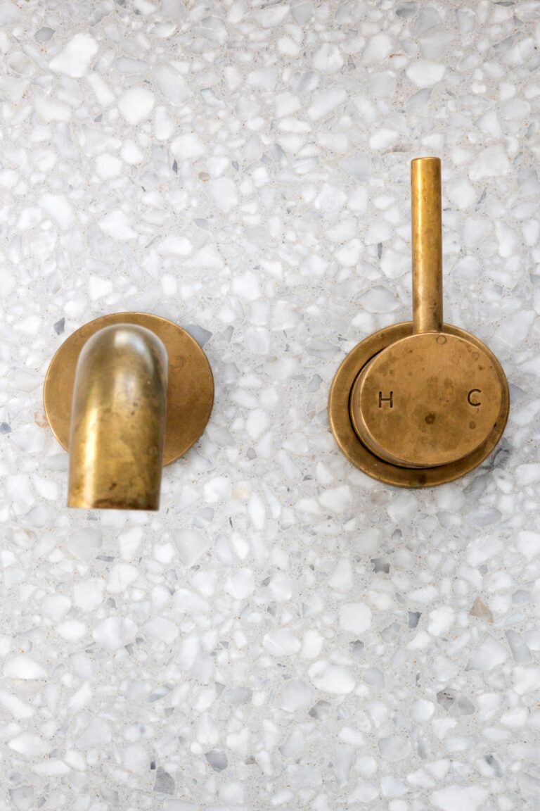 Detail of a luxury bathroom from a home renovation in Sunset Vancouver featuring a wall-mounted aged brass faucet and temperature control lever against white terrazzo stone tiling.