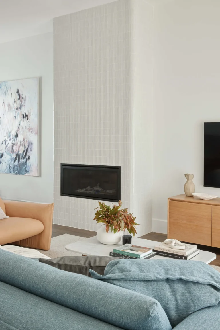 A modern living room from a home renovation in Victoria–Fraserview featuring a floor-to-ceiling white tiled fireplace, a minimalist oak media console, and contemporary blue and tan furniture.