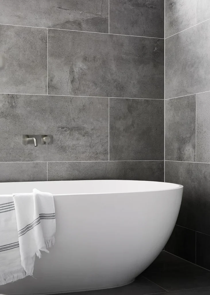A modern bathroom renovation in North Vancouver BC by Quay Construction featuring a white freestanding soaking tub against a large-format charcoal grey stone tile wall with a minimalist wall-mounted faucet.