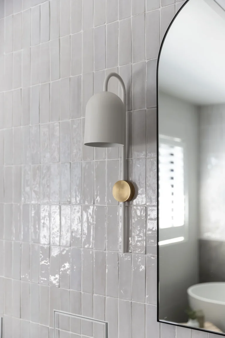 Modern bathroom renovation in Oakridge Vancouver featuring white vertical stacked tiles and a minimalist wall sconce with gold accents next to an arched mirror.