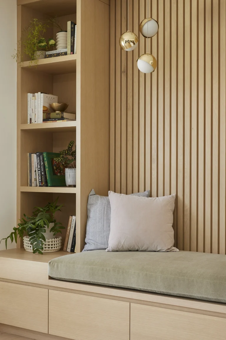 A custom-built reading nook from a home renovation in Yaletown featuring an oak slat accent wall, integrated bookshelf, and a sage green cushioned bench with gold globe pendant lights.
