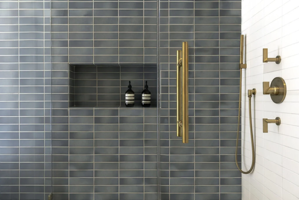 A luxury walk-in shower from a home renovation in Downtown Vancouver featuring blue vertical stacked tiles, a recessed wall niche, and premium brushed gold hardware including a handheld sprayer.