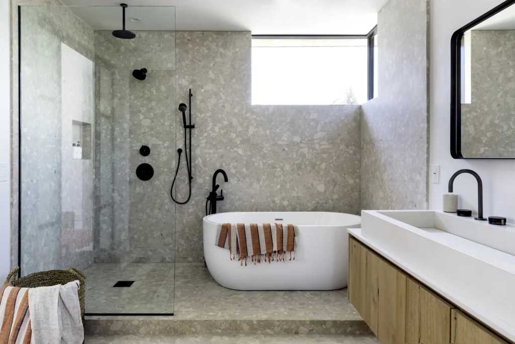 Modern bathroom from a home renovation in Hastings–Sunrise featuring a curbless glass shower, freestanding white soaking tub, and terrazzo stone walls with matte black fixtures.