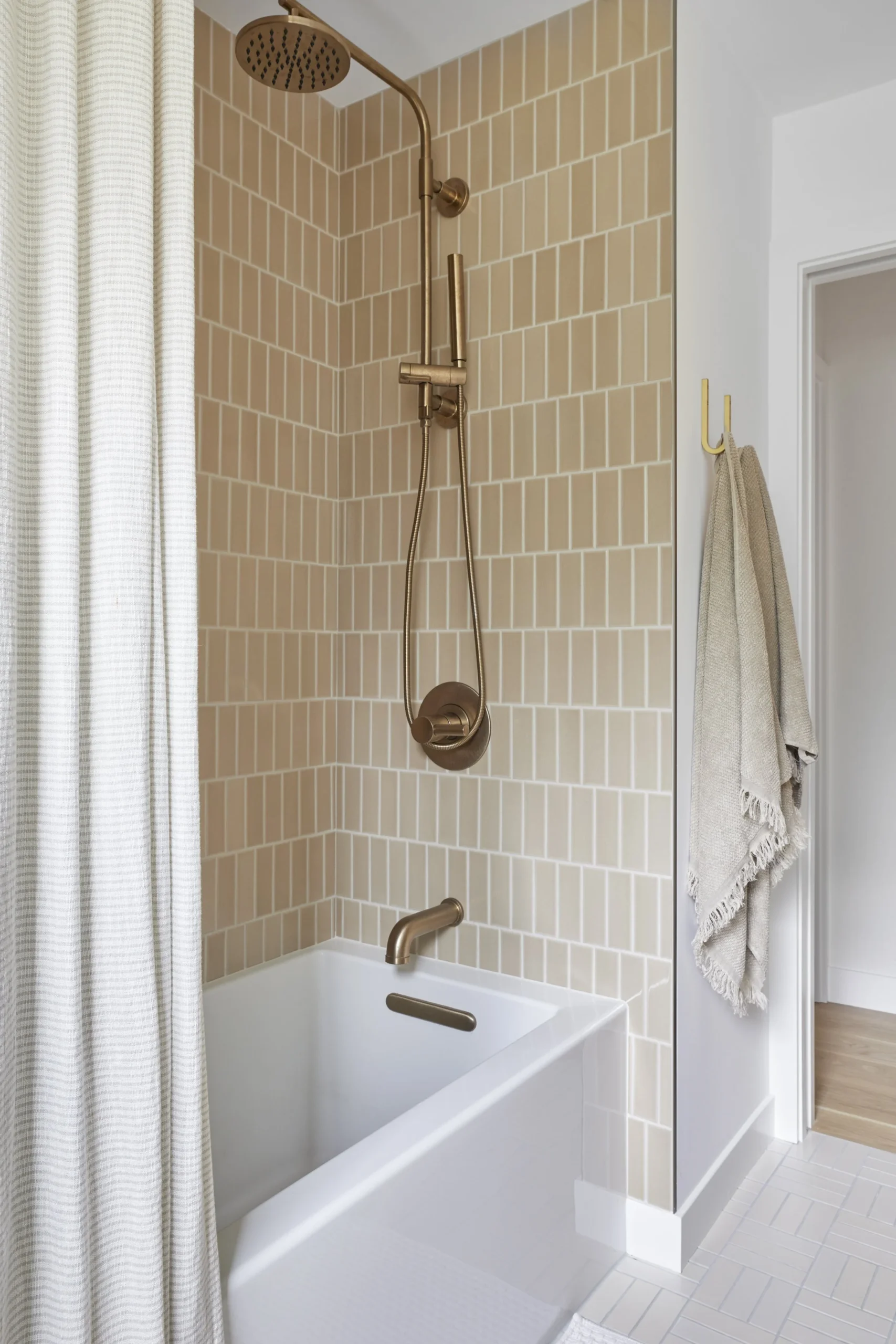 Modern bathroom renovation in Metrotown by Quay Construction featuring vertical stacked beige subway tiles, brushed gold shower fixtures, and a white soaking tub.
