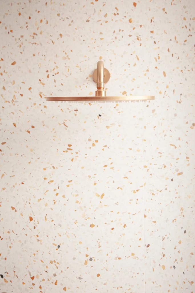 A luxury walk-in shower detail by Vancouver renovation company Quay Construction featuring a large brushed copper rain shower head against a warm-toned terrazzo tile wall.
