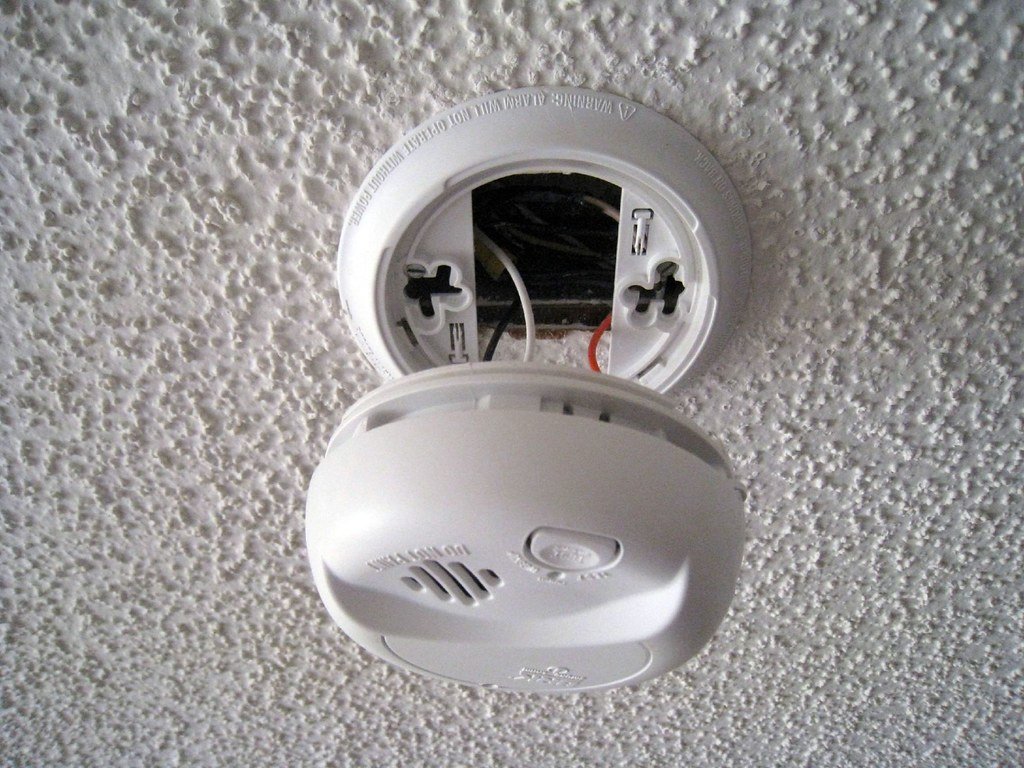 A hard-wired smoke alarm opened from its ceiling base, showing the wiring connector