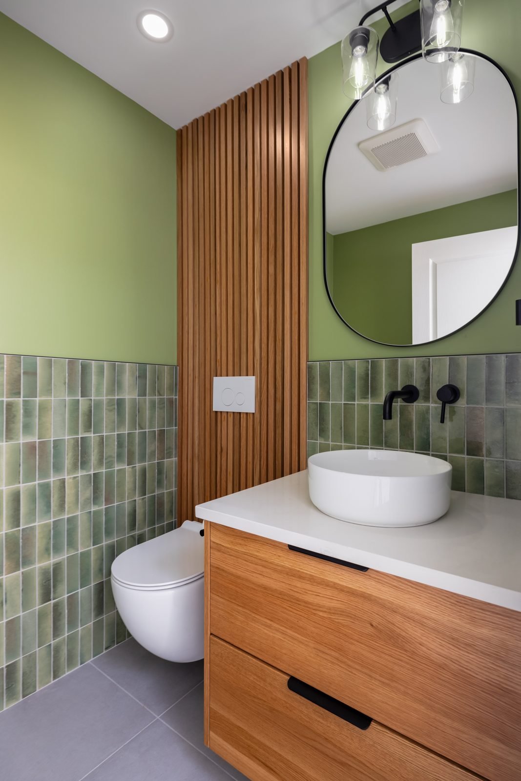 Powder room with a wood slat feature wall, green tile wainscot, vessel sink and oval mirror in North Vancouver