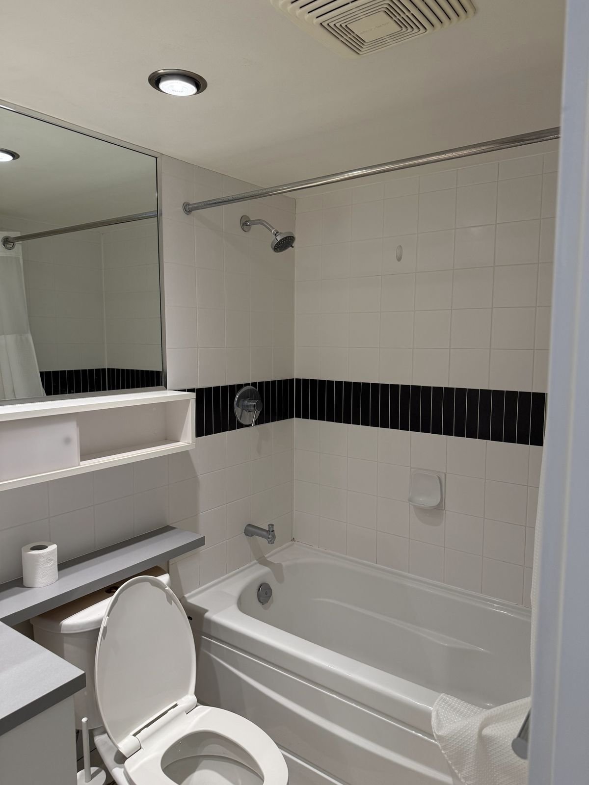 Before: Yaletown condo bathroom with a dated tub, white wall tile, black accent band and shower curtain